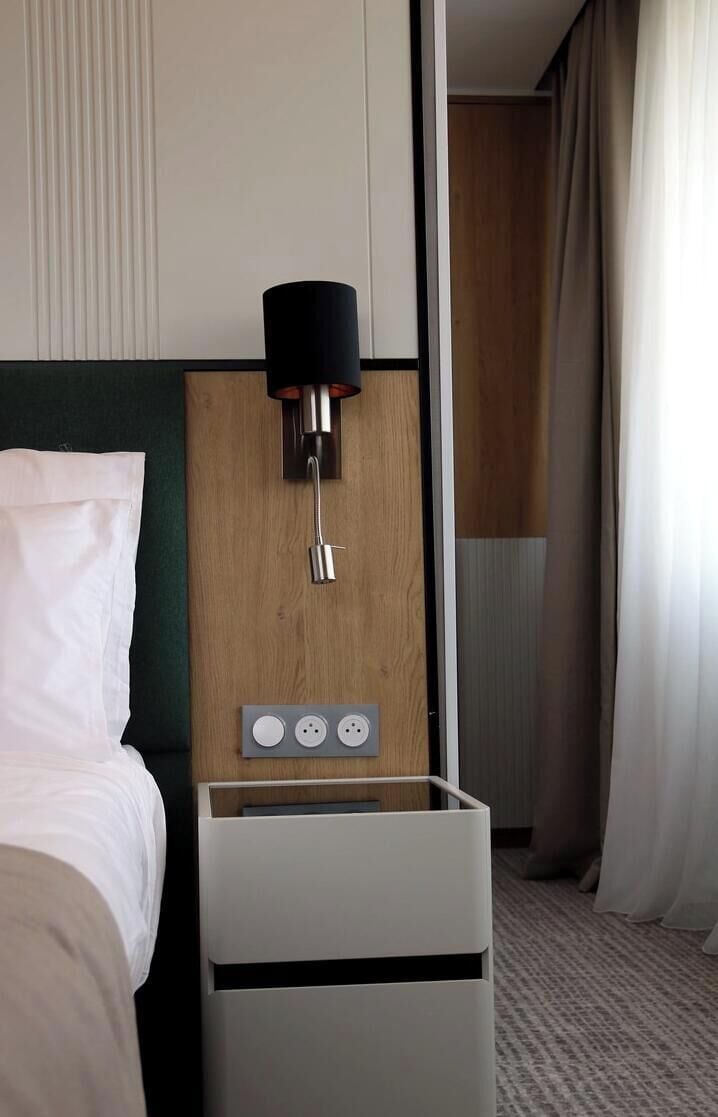 A hotel room with a bed, a wall-mounted lamp, and a thermostat control panel.