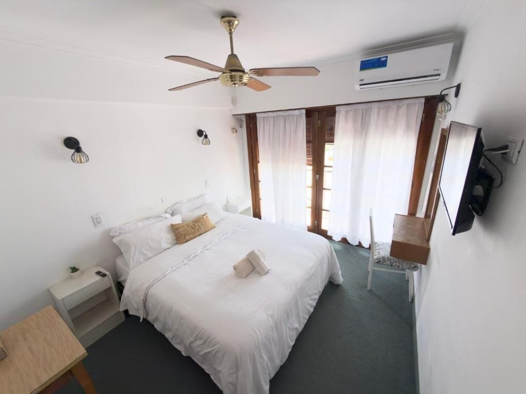 Een hotelkamer met een bed, een plafondventilator, een airconditioningapparaat, een nachtkastje en een aan de muur bevestigde televisie.