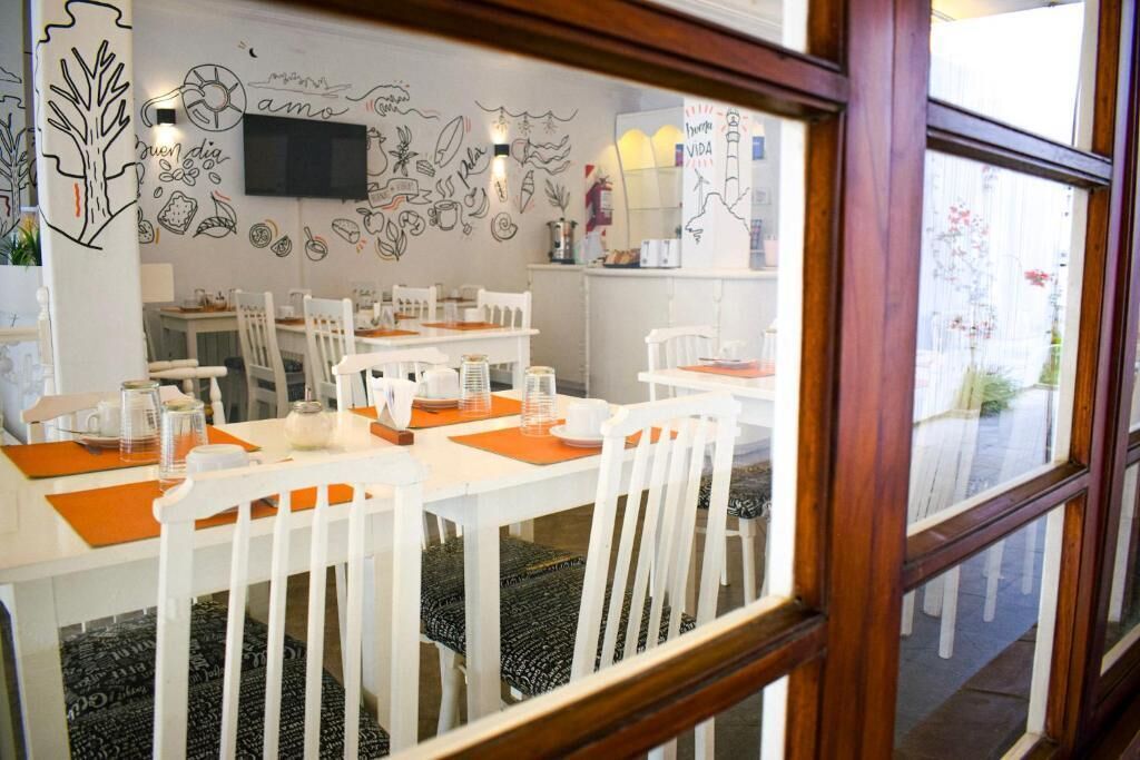 Un comedor con mesas y sillas blancas, un televisor montado en la pared y una ventana con vistas al exterior.