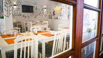 Un comedor con mesas y sillas blancas, un televisor montado en la pared y una ventana con vistas al exterior.