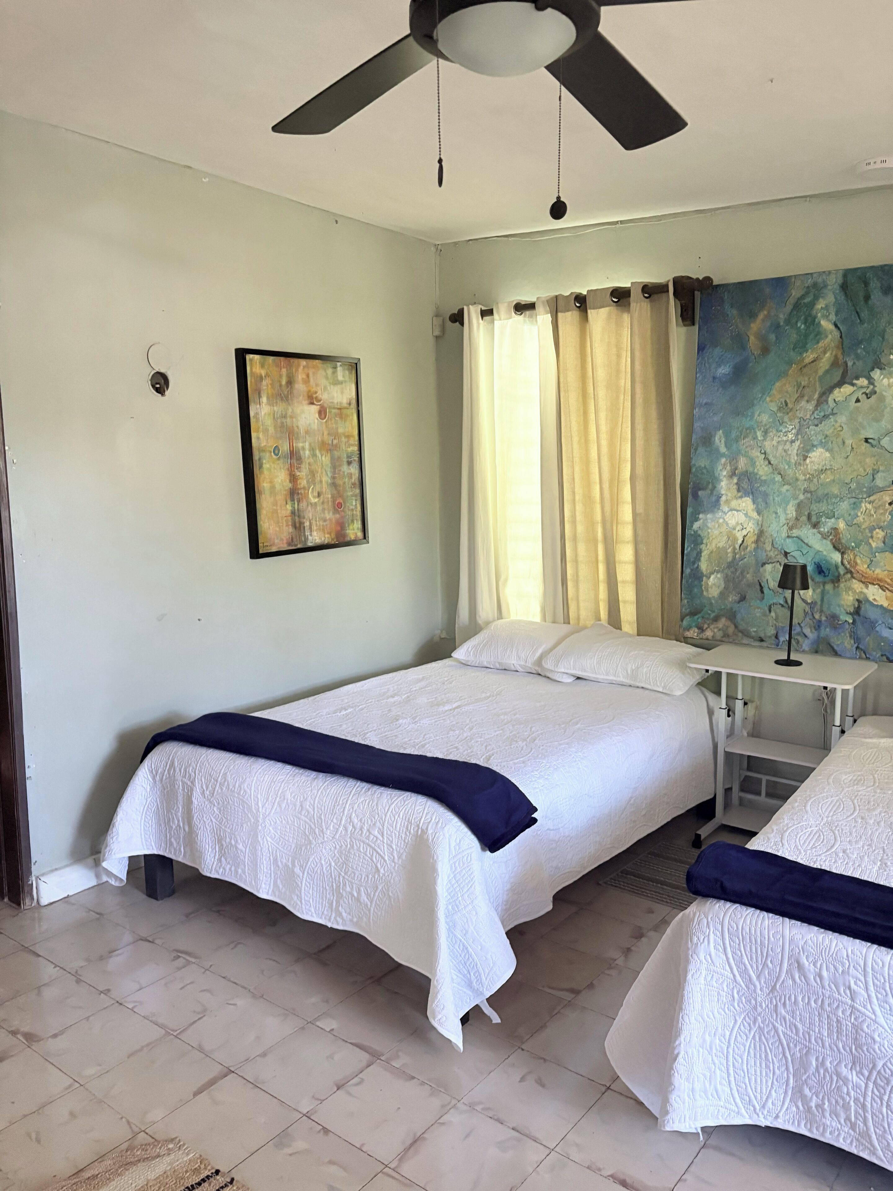 Une chambre d’hôtel avec deux lits, un ventilateur de plafond, un tableau et une grande fenêtre avec des rideaux.