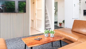 Un salón moderno con un televisor montado en la pared, una mesa de centro de madera con controles remotos y plantas en macetas, un sofá de cuero marrón y un armario blanco.