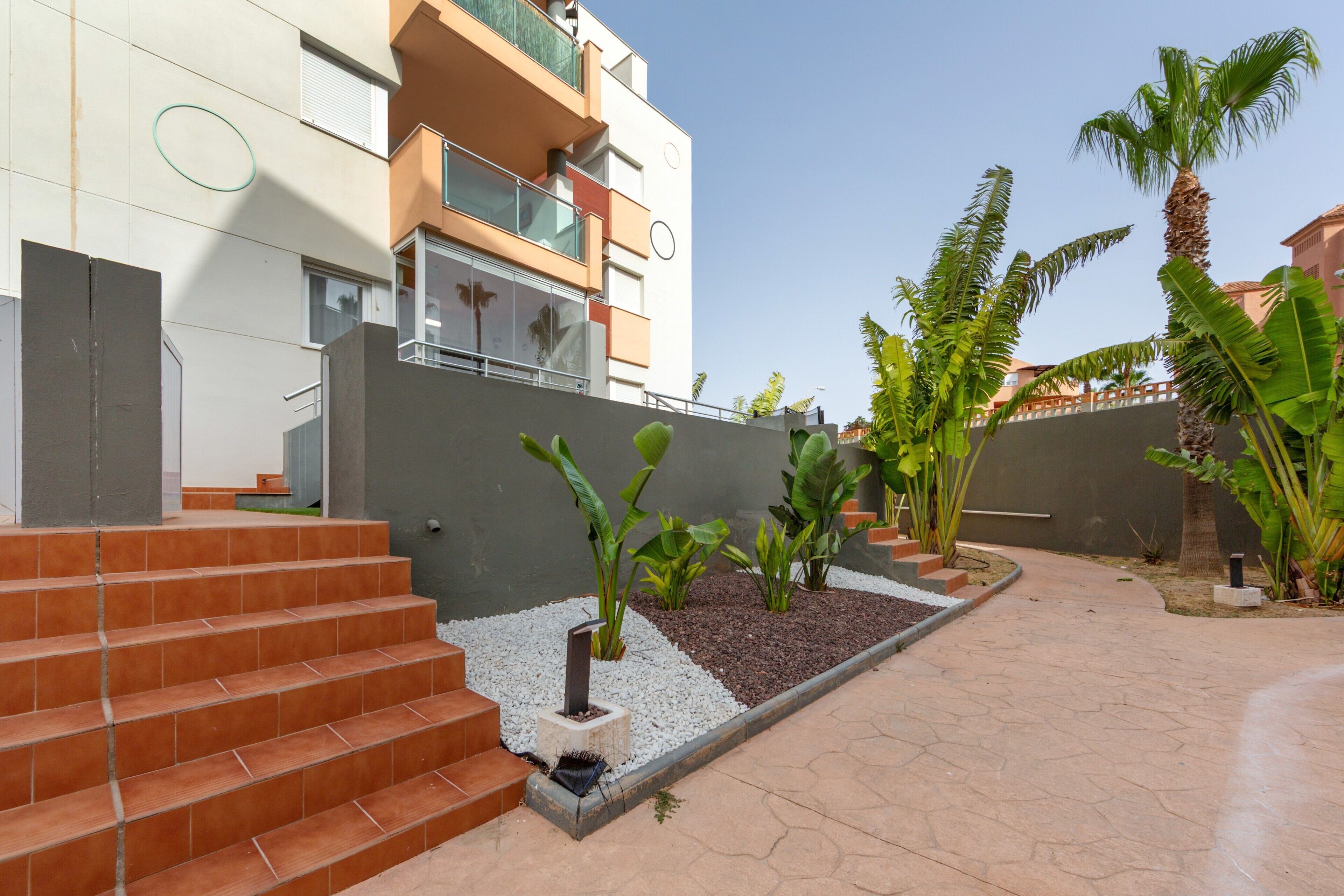 Un edificio moderno con una escalera que conduce a un balcón, rodeado por un jardín con palmeras y un camino pavimentado.