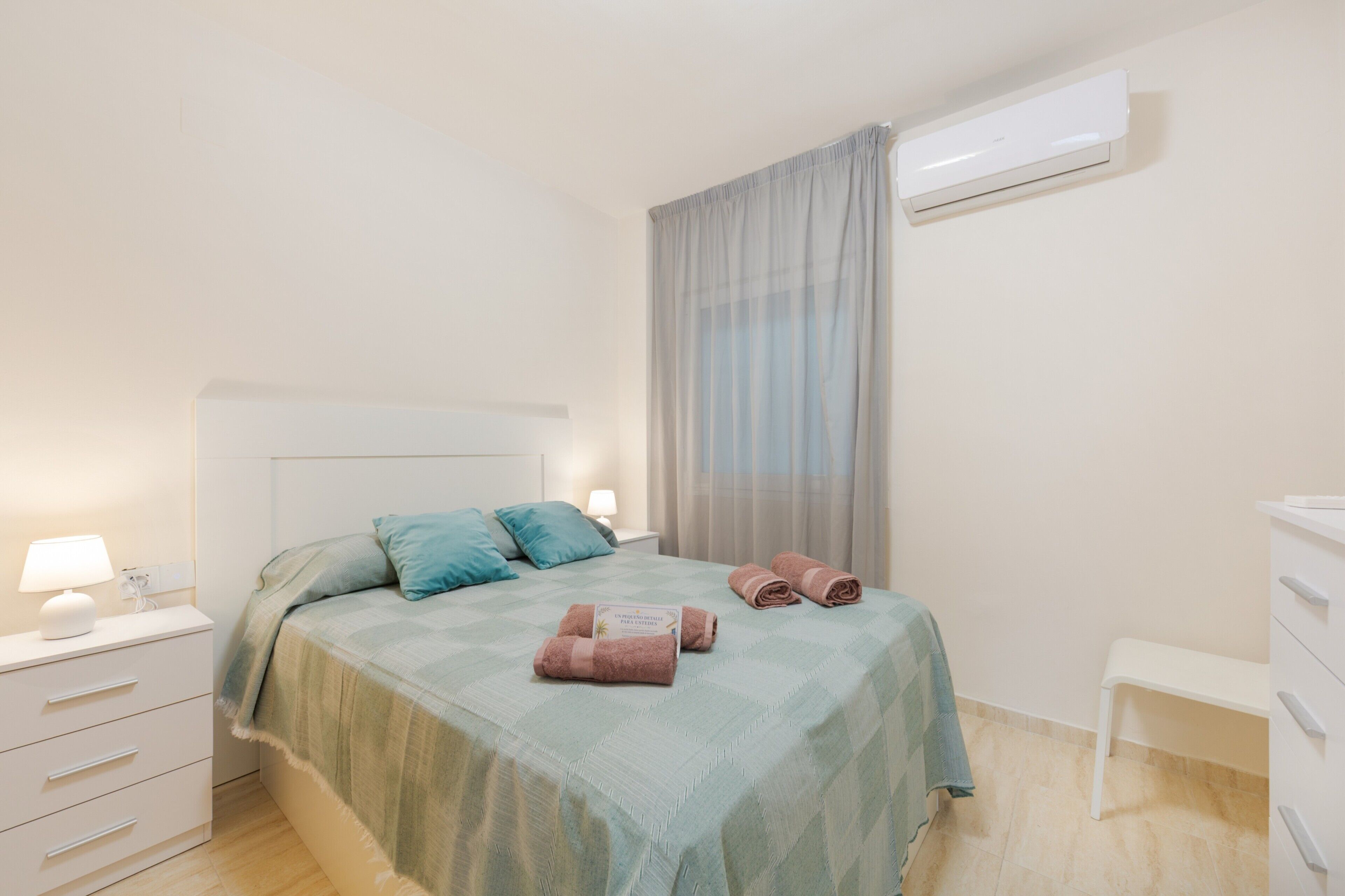 Een netjes opgemaakt bed met een blauwgroene sprei, een nachtkastje met een lamp en een witte dressoir.