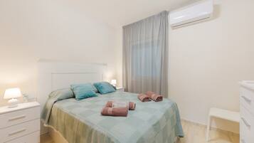 Een netjes opgemaakt bed met een blauwgroene sprei, een nachtkastje met een lamp en een witte dressoir.