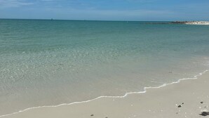 Uma praia com águas cristalinas, areal arenoso e um afloramento rochoso ao longe.