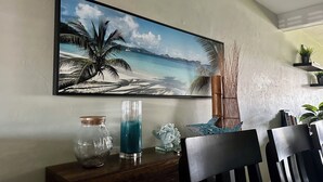 Uma área de jantar com uma pintura de uma cena de praia, um vaso de vidro, um recipiente de vidro azul e uma cesta trançada sobre uma mesa de madeira.