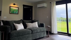 A living room with a sofa, a bench, a painting, an air conditioning unit, and a large window with a view. - Charming 3-bedroom House on a Cherry Orchard (Suttons Bay)