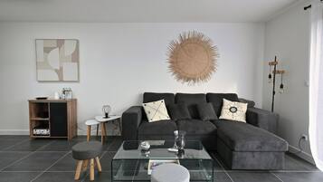 Ett modernt vardagsrum med en hörnsoffa, ett soffbord i glas, ett litet sidobord och en dekorativ väggdekoration.