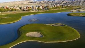 En golfbane med bunkers og et vandhazard, omgivet af bygninger og bjerge.