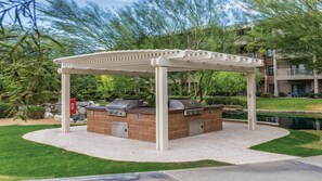 Et udendørs grillområde med pergola, stenbordplade og en lille dam i baggrunden.