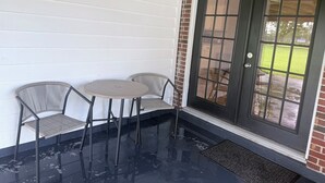 A covered patio area with two wicker chairs and a small round table, a black door with glass panels, and a wet floor. - Home Sweet Home Nearby Beach & MS Gulf Coast! (Grand Bay)