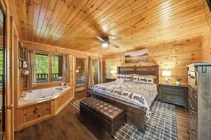 A wooden cabin bedroom with a large bed, a separate bathroom with a bathtub, a ceiling fan, and a view of the outdoors. - Peaceful Log Cabin Retreat with Hot Tub (Sevierville)