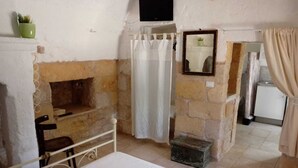 A room with a stone fireplace, a bed, a small table, a mirror, and a television mounted on the wall. - LA TORRETTA SHORT-TERM RENTALS (MONOPOLI)