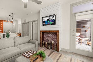 A modern living room with a flat-screen TV mounted above a brick fireplace, a grey sofa, and a wicker coffee table with a remote control and decorative items. - Downtown Suite on San Antonio Riverwalk Walkable to the Alamo (San Antonio)