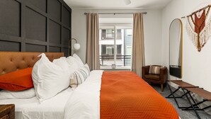 Habitación de hotel con cama, una silla, un banco, ventilador de techo y un espejo grande, con vista al balcón exterior.