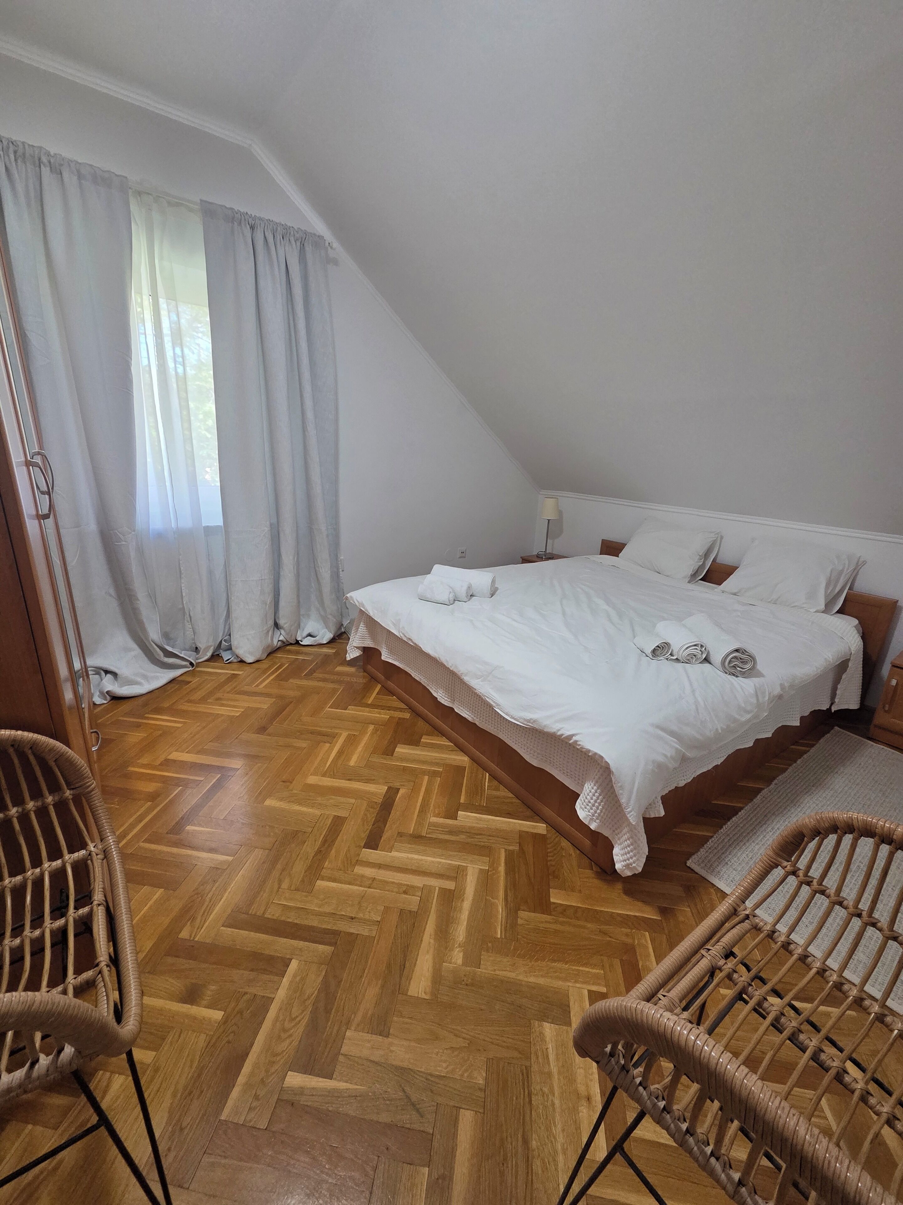 A bedroom with a bed, two wicker chairs, a wooden herringbone floor, and a window with curtains.