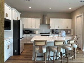 A modern kitchen with white cabinetry, a central island with bar stools, and built-in appliances. - 4BR Home: Theme Parks & Huntington Beach Nearby (Garden Grove)
