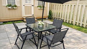 A patio with a glass table and four chairs, a black umbrella, and a wooden fence in the background.