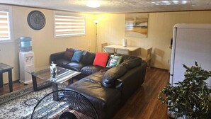 A living room with a leather sofa, a glass coffee table, a water dispenser, a fan, and a refrigerator.