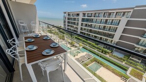 Ein Balkon mit Holztisch und weißen Stühlen, mit Blick auf einen Swimmingpool und ein Gebäude mit mehreren Balkonen.