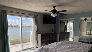Um quarto com cama, penteadeira, ventoinha no teto, TV e uma grande janela com vista para um lago.
