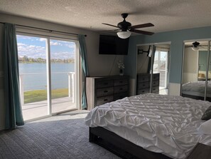 Ein Schlafzimmer mit Bett, Kommode, Deckenventilator, Fernseher und einem großen Fenster mit Seeblick.