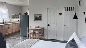 A bedroom with a bed, bedside table, and a small desk with chairs. A kitchen with cabinets and a refrigerator is visible in the background. - Lovely studio in charming Arcata perfect for your stay (Arcata)