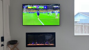 A room with a television mounted on the wall displaying a sports event, a fireplace below it, and a wooden rack to the left. - Lovely studio in charming Arcata perfect for your stay (Arcata)