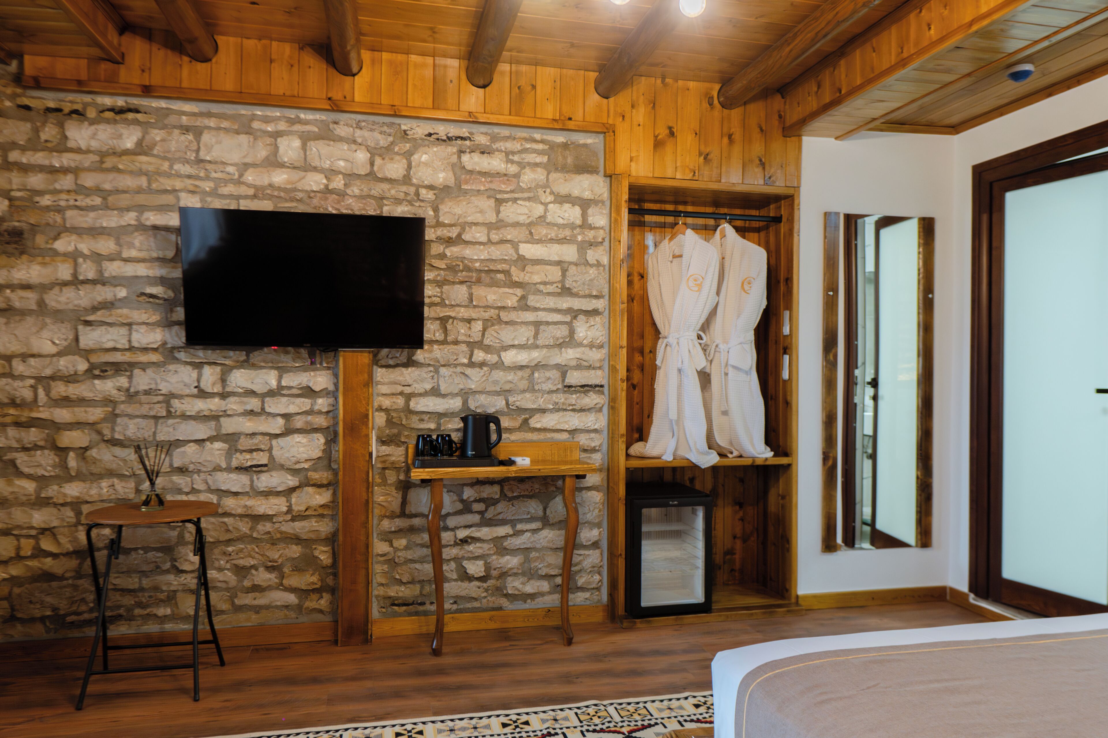 Ein Zimmer mit Steinwand, Holzboden, einem Flachbildfernseher, einem kleinen Tisch mit Kaffeekocher und einem Kleiderschrank mit Bademänteln.