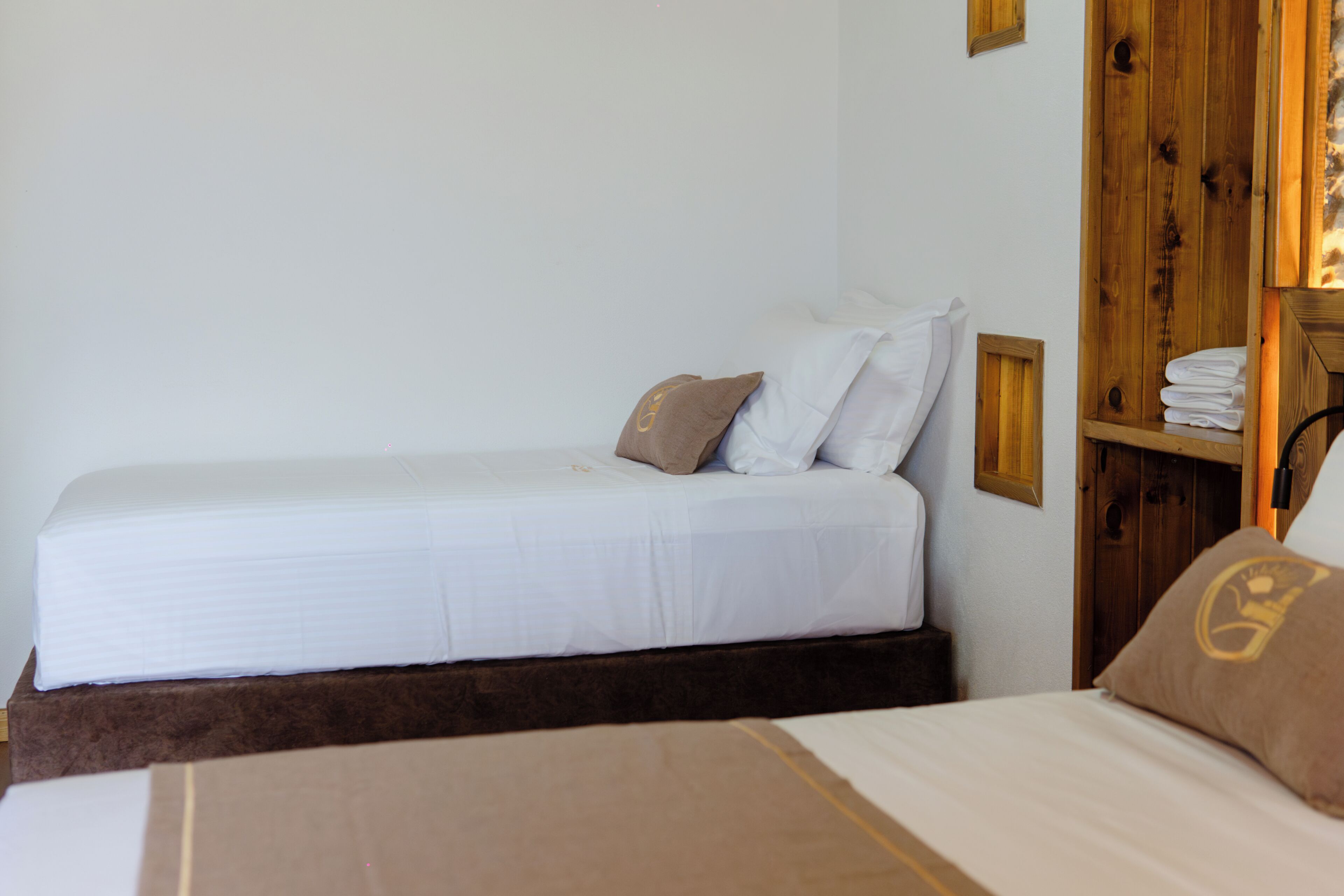 Una cama individual con colchón blanco y dos almohadas, cabecera de madera y un pequeño estante con toallas dobladas.