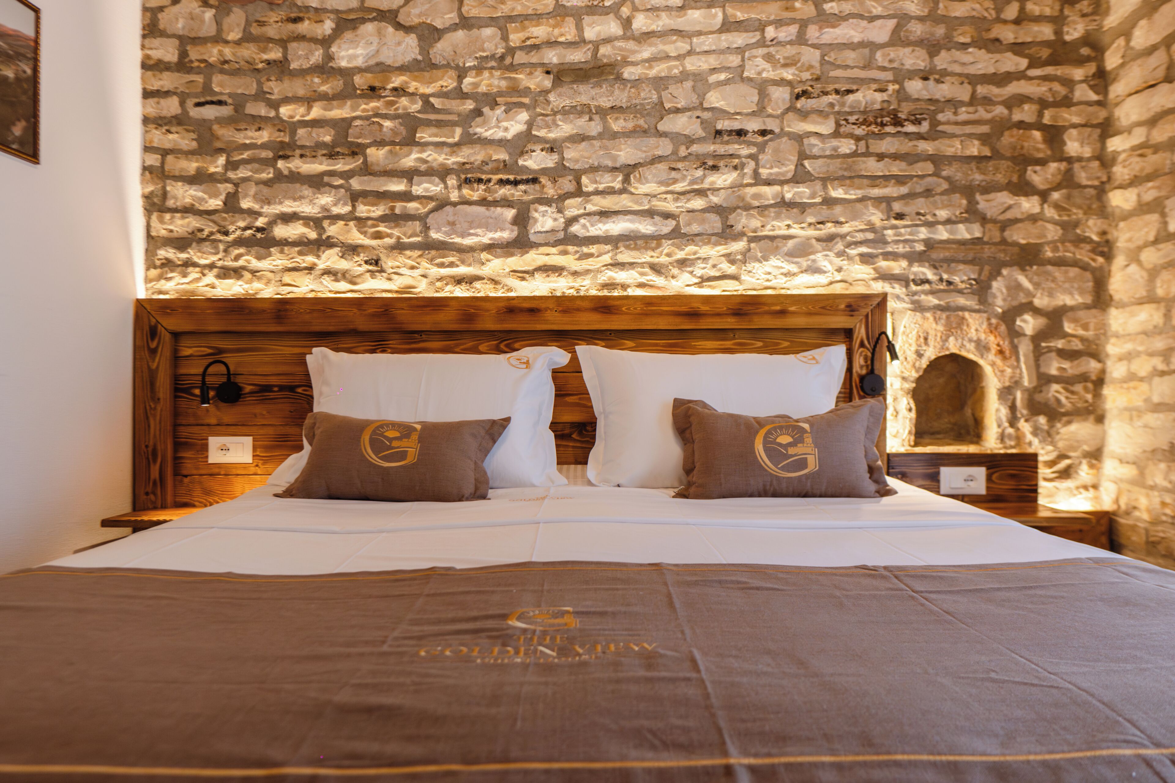 A bed with a stone wall backdrop, wooden headboard, and two pillows with a logo.