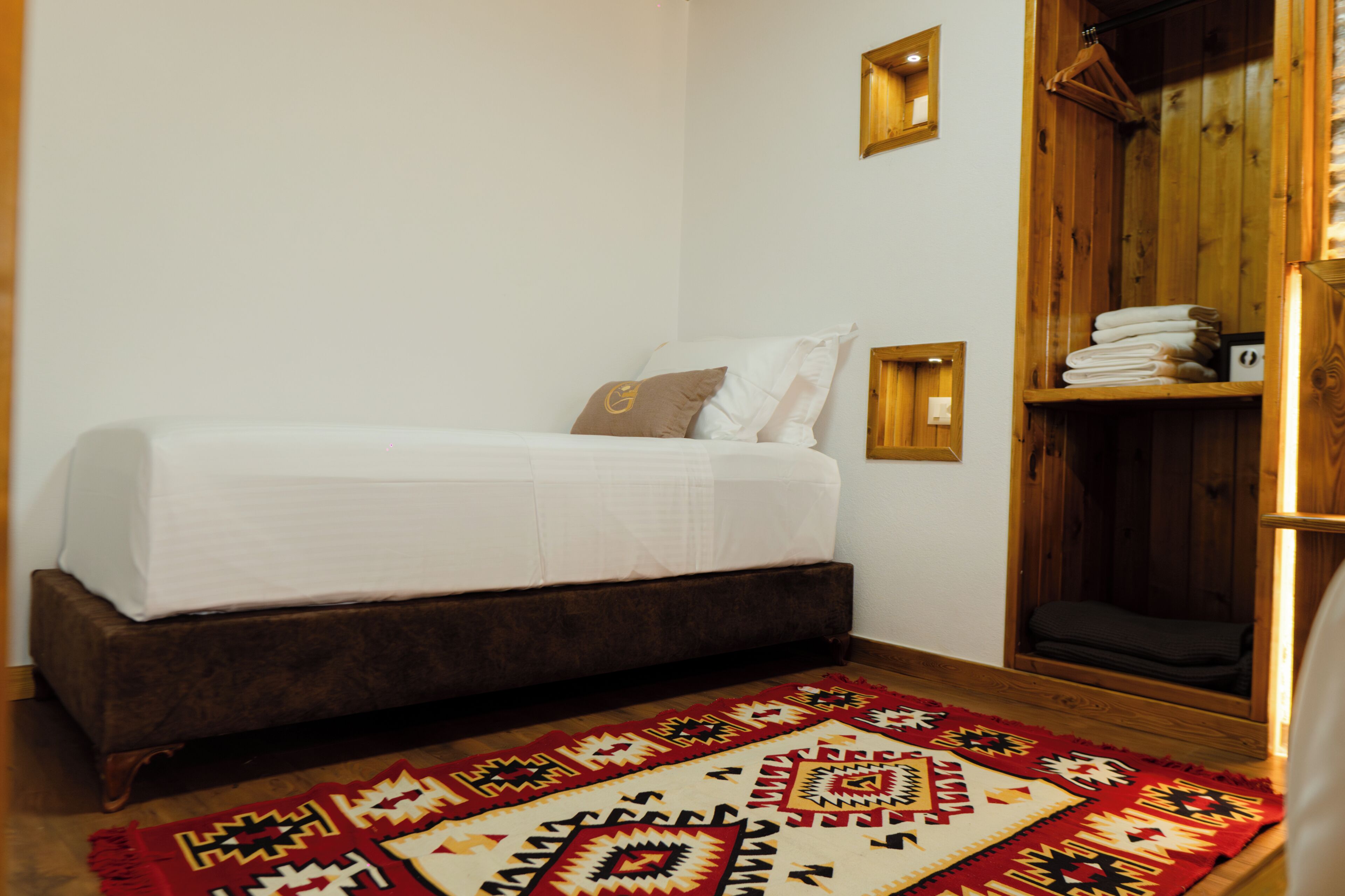 A room with a bed, a wooden wardrobe, a patterned rug, and a wall-mounted light fixture.