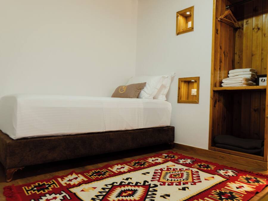 A room with a bed, a wooden wardrobe, a patterned rug, and a wall-mounted light fixture.
