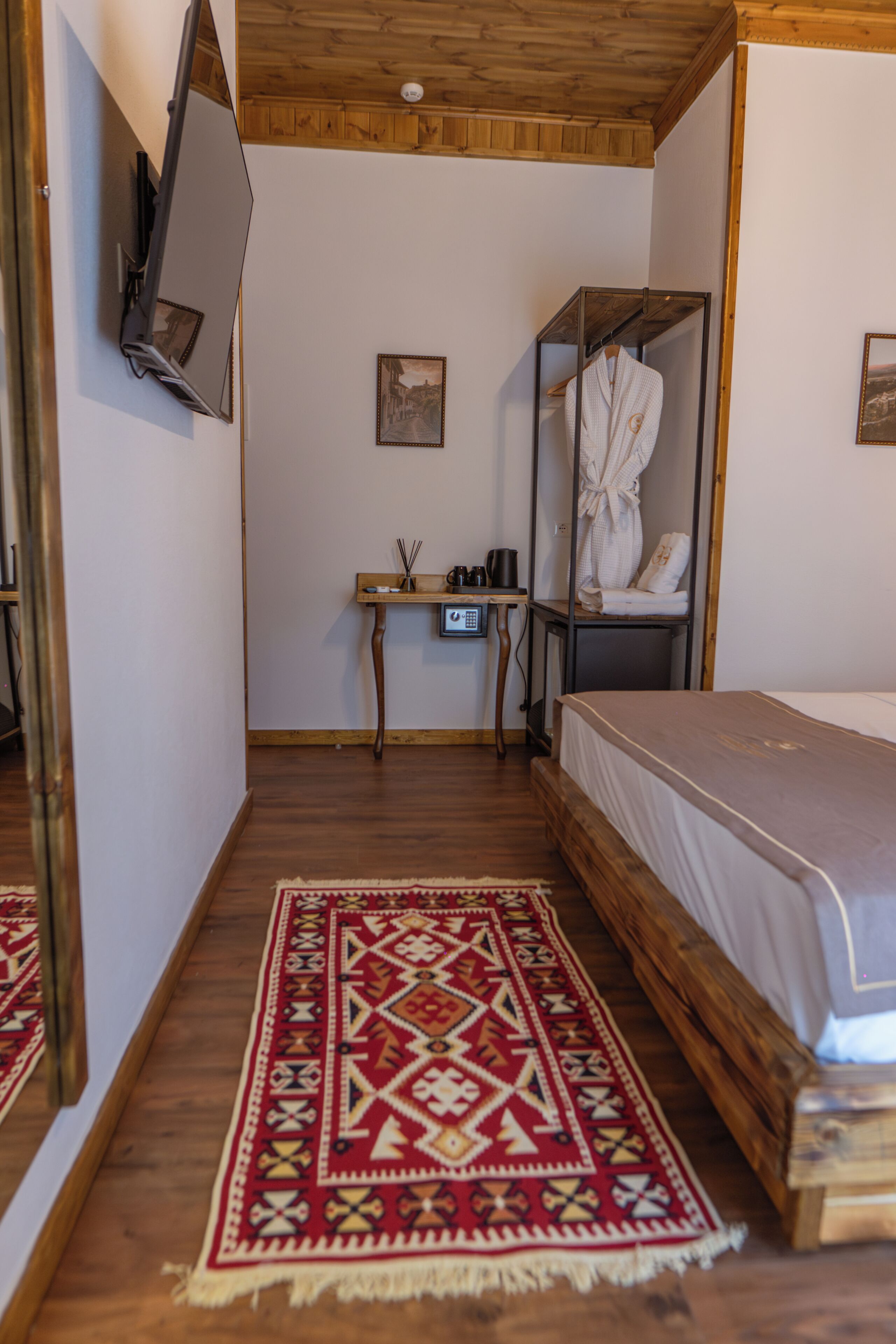 Un dormitorio con techo de madera, una cama, un televisor montado en la pared, un escritorio con una silla y una alfombra roja estampada.