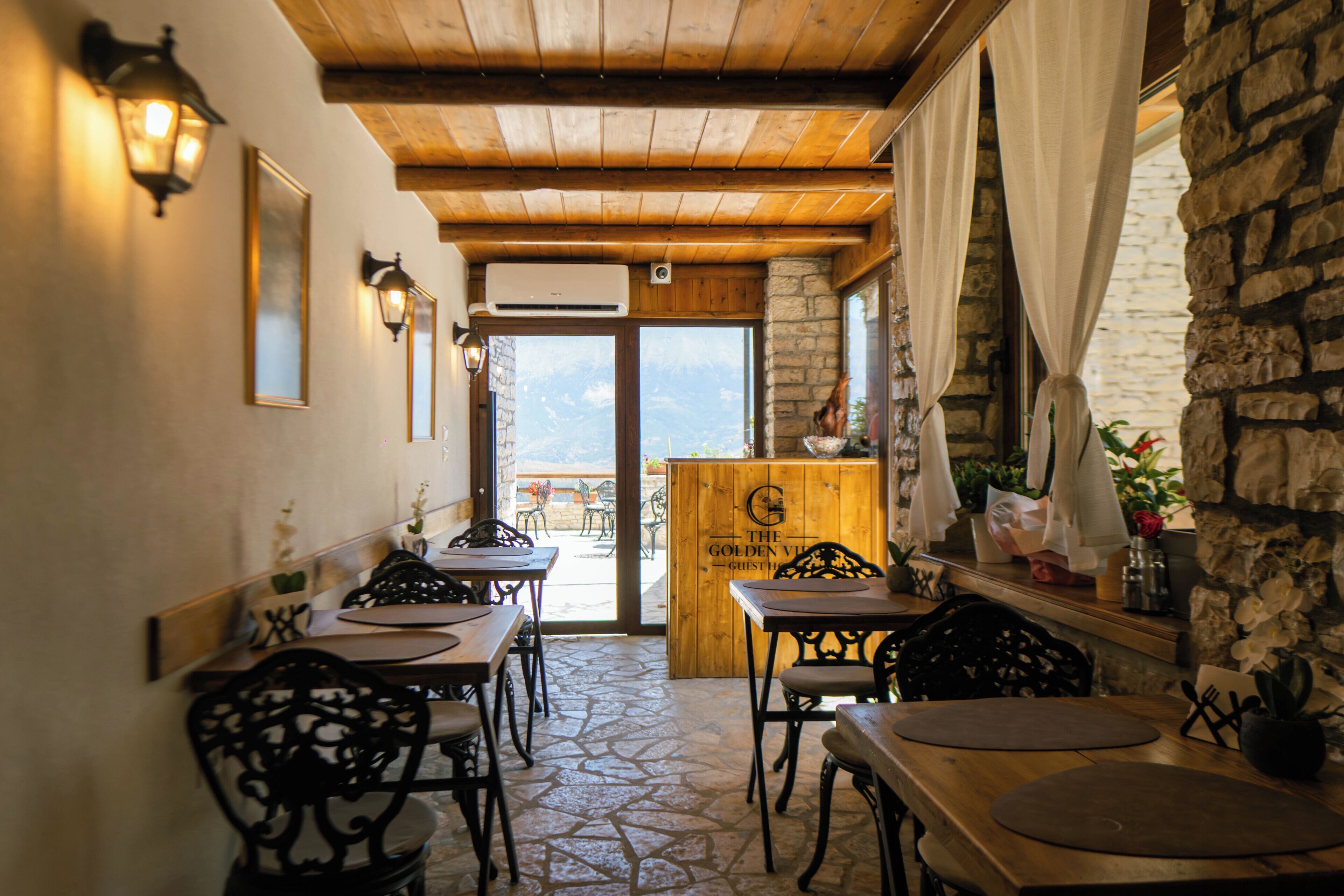 Een restaurantinterieur met houten tafels en stoelen, stenen vloeren en een uitzicht door de deur.