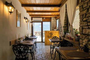 L’intérieur d’un restaurant avec des tables et des chaises en bois, un sol en pierre et une vue sur l’extérieur à travers la porte.