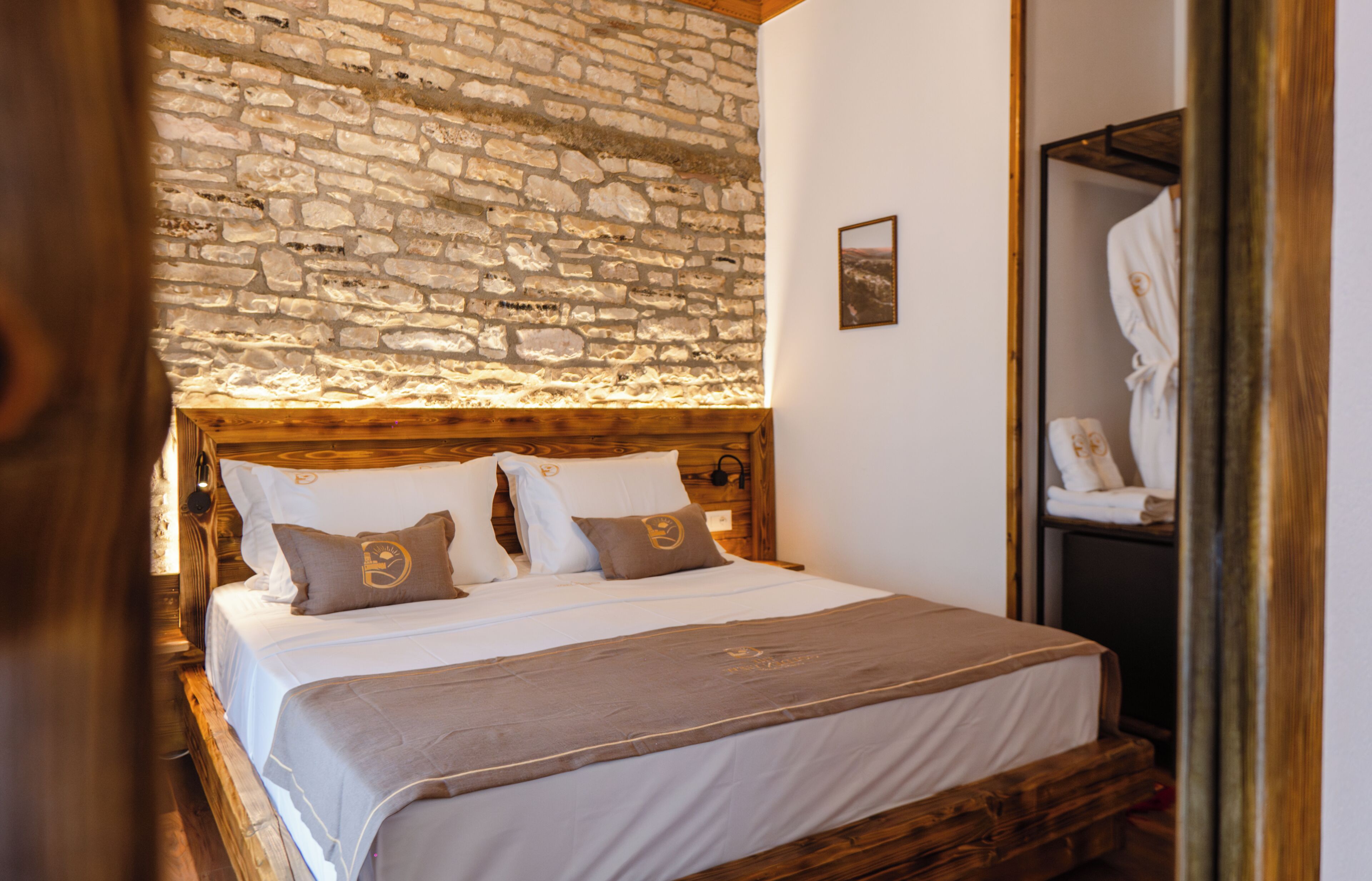 Un dormitorio con pared de piedra, cabecera de madera, cama con ropa blanca y dos almohadas, y una mesita de noche de madera.