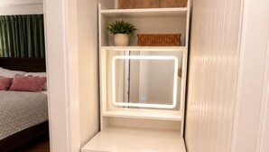 Una camera d'angolo con un letto, un comodino bianco con cassetti, uno specchio e un piccolo ripiano con una pianta e un cesto.