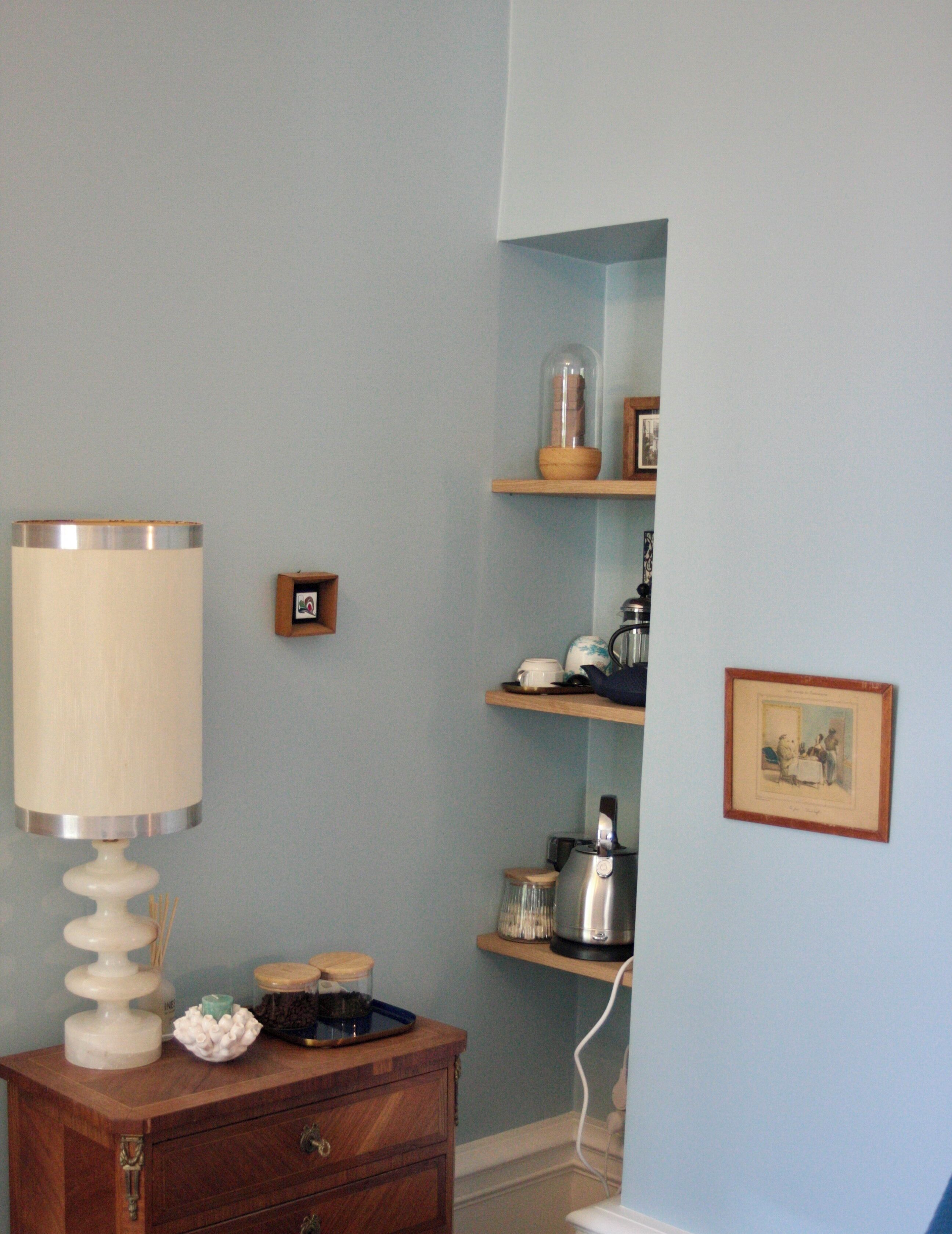 A room with a wooden dresser, a lamp, a shelf with a picture frame, and a wall-mounted picture.