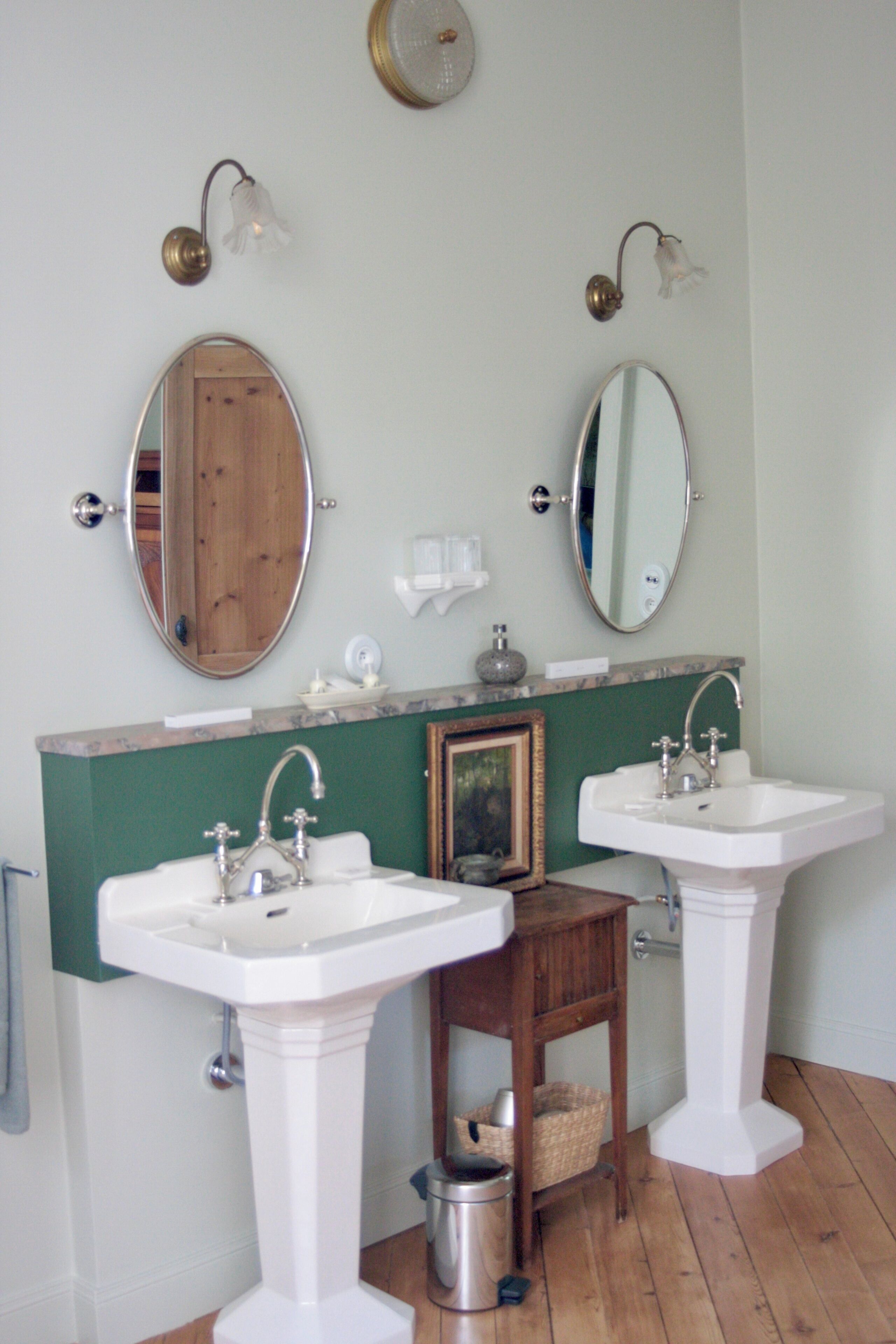 Un baño con dos lavamanos blancos de pedestal, un mueble de tocador de madera y dos espejos montados en la pared.