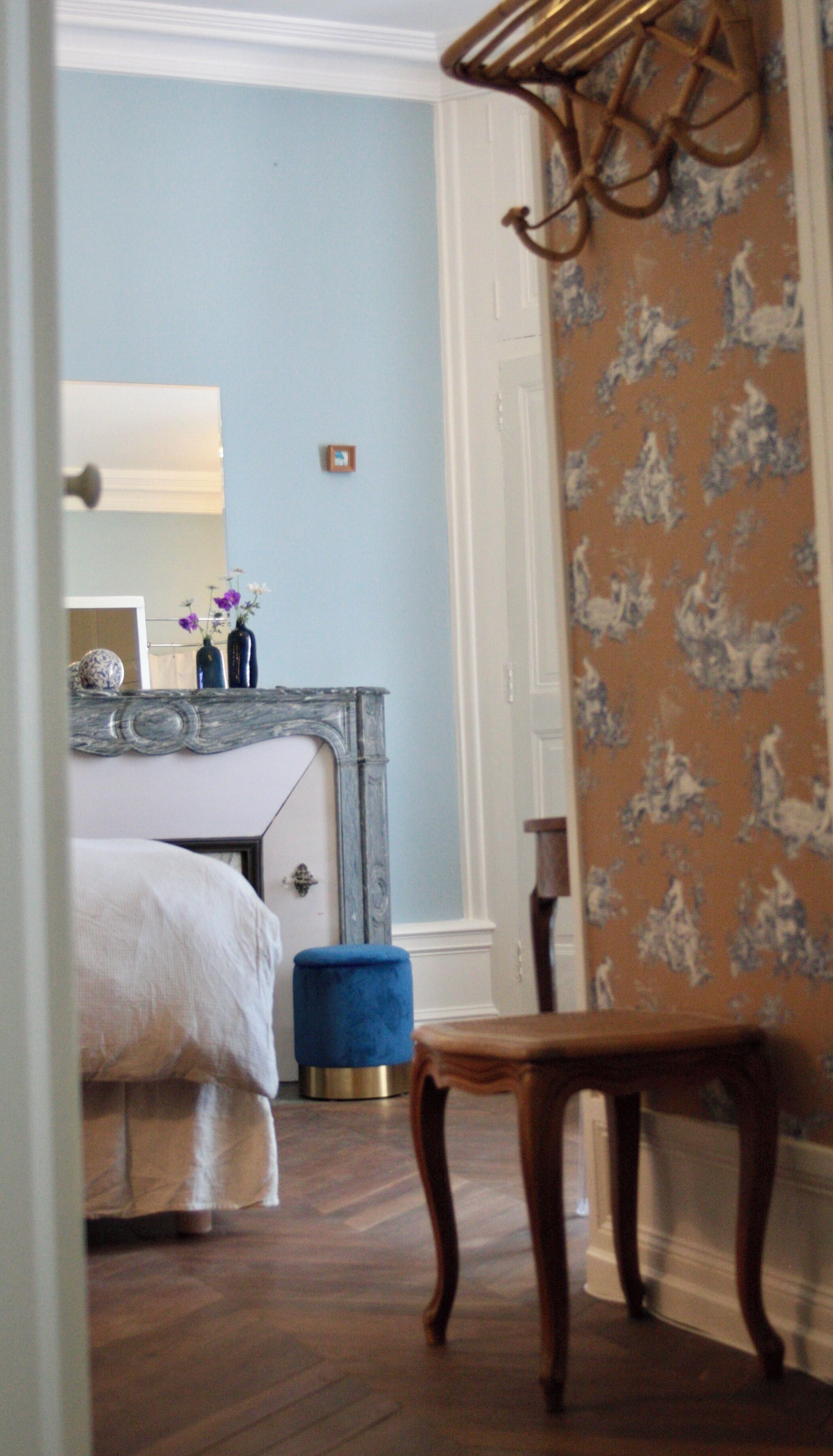 A bedroom with a bed, a blue ottoman, a wooden chair, a mirror, and a door leading to another room.