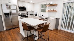 Una cocina moderna con armarios blancos, electrodomésticos de acero inoxidable, una isla central con taburetes y un ventanal con persianas.