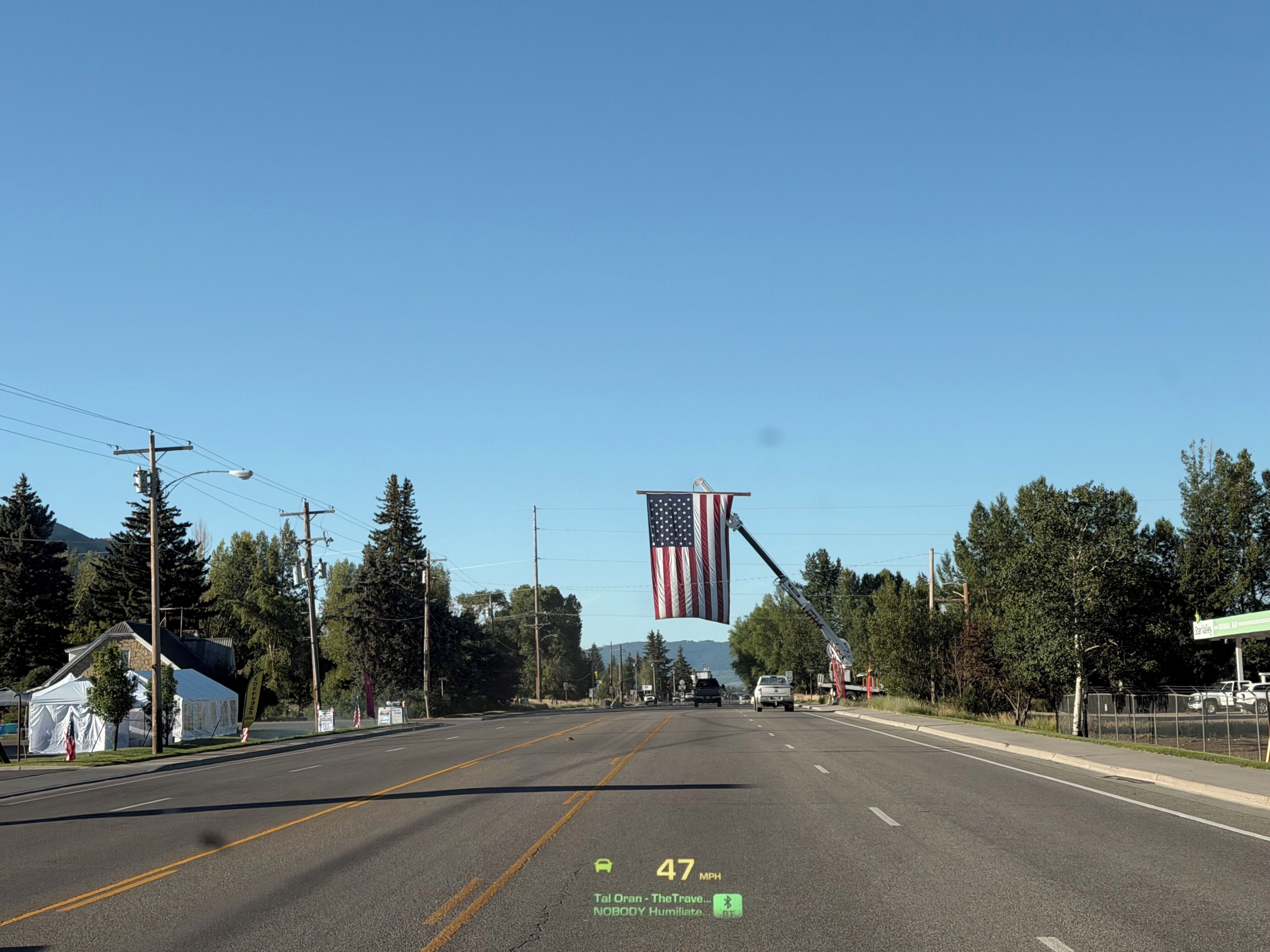 Carretera con un cartel de límite de velocidad de 76 kilómetros por hora, una gran bandera estadounidense y un cielo azul despejado.