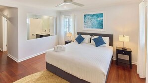 A bedroom with a bed, bedside tables, a ceiling fan, a window with curtains, a wall-mounted mirror, and a painting.