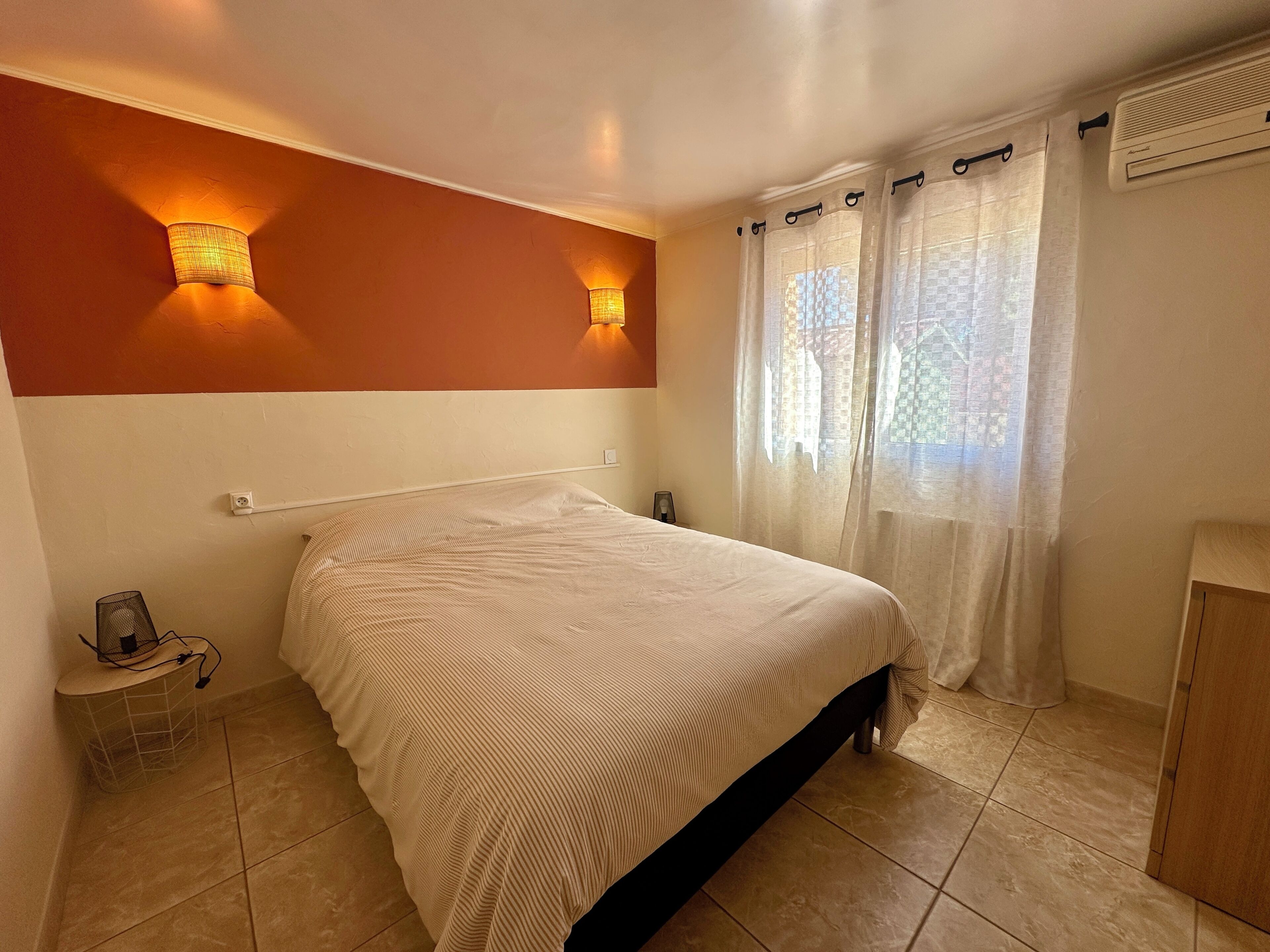 Une chambre à coucher comprenant un lit, deux lampes fixées au mur, une fenêtre avec des rideaux, une table de chevet et un thermostat.