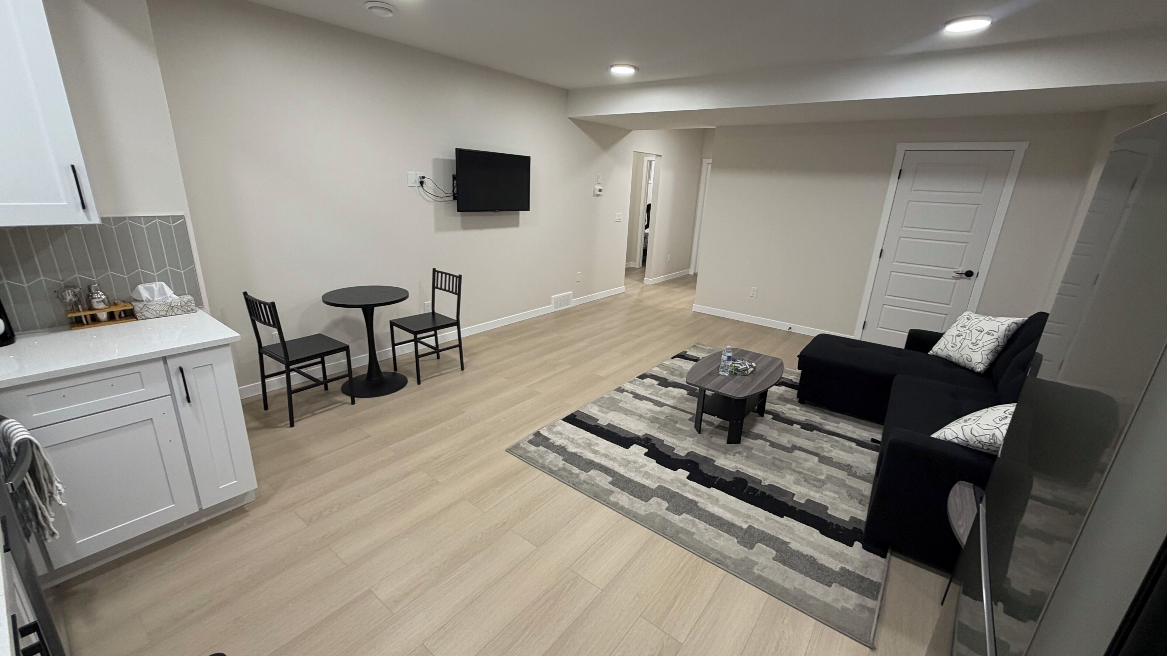 Een moderne woonkamer met een bank, salontafel en stoelen. Er is een keukenhoek met kastjes en een aanrecht. Een flatscreen televisie is aan de wand bevestigd.