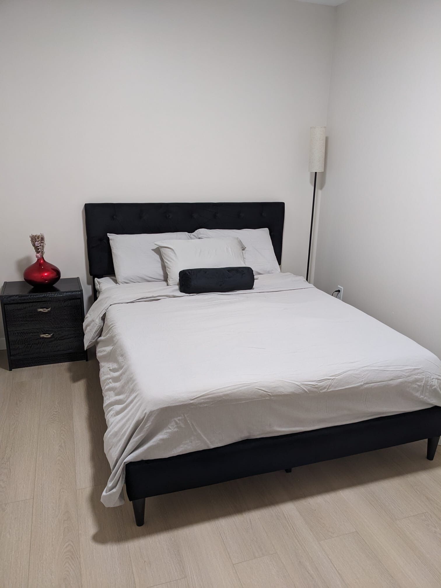 Una cama con ropa de cama blanca, cabecera negra y mesita de noche con un florero rojo.