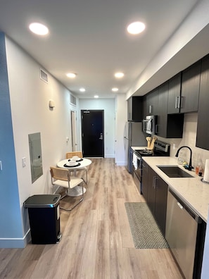 A modern kitchen with dark cabinets, a stainless steel refrigerator, a microwave, and a sink. There is a dining area with a table and chairs.