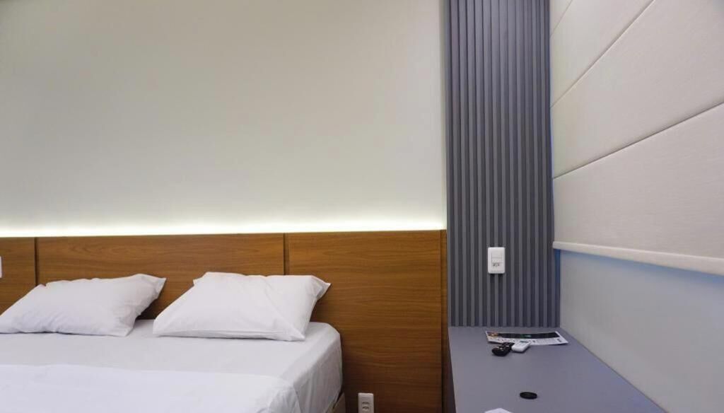 Ein Hotelzimmer mit Holz-Kopfteil, einem Bett mit weißen Bettwäsche, einem Nachttisch mit Fernbedienung und einer Wandleuchte.
